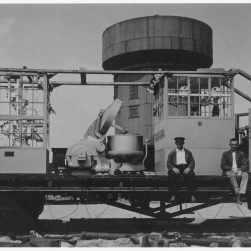 Platte goederenwagon met een bakkerijmachine voor bakkerij van Steenland op het spoorwegemplacement van Gouda; daarachter de watertoren voor de waterinname van stoomlocomotieven; geheel rechts (met hoed): Theo Gerardus Steenland sr.; links daarvan: Theo Geradus Steenland jr.; circa 1930 (SAMH 0440. 25844)